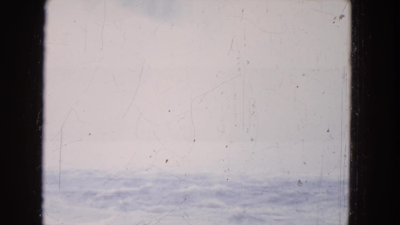 1964 8mm travel-film frame showing a rippled water band beneath pale sky, with scratches and slight exposure variation.