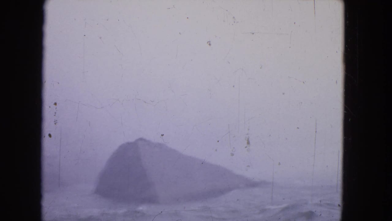 Dark rock silhouette surrounded by mist and water on a scratched purple-tinted frame.