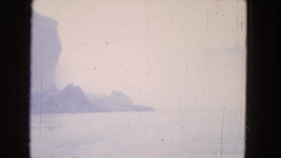 Niagara Falls appears through haze in a 1964 8mm frame, with a darker cliff silhouette and calm water below.