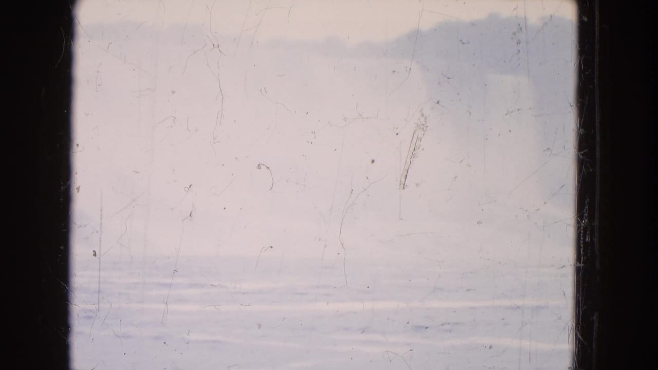 1964 8mm frame of a hazy waterside view with pale sky, muted water texture, and dark vertical borders.