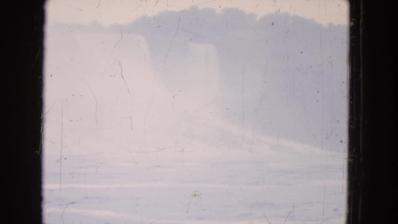 1964 8mm home-movie frame with a hazy landform, pale sky, and softly rippled water across the lower view.