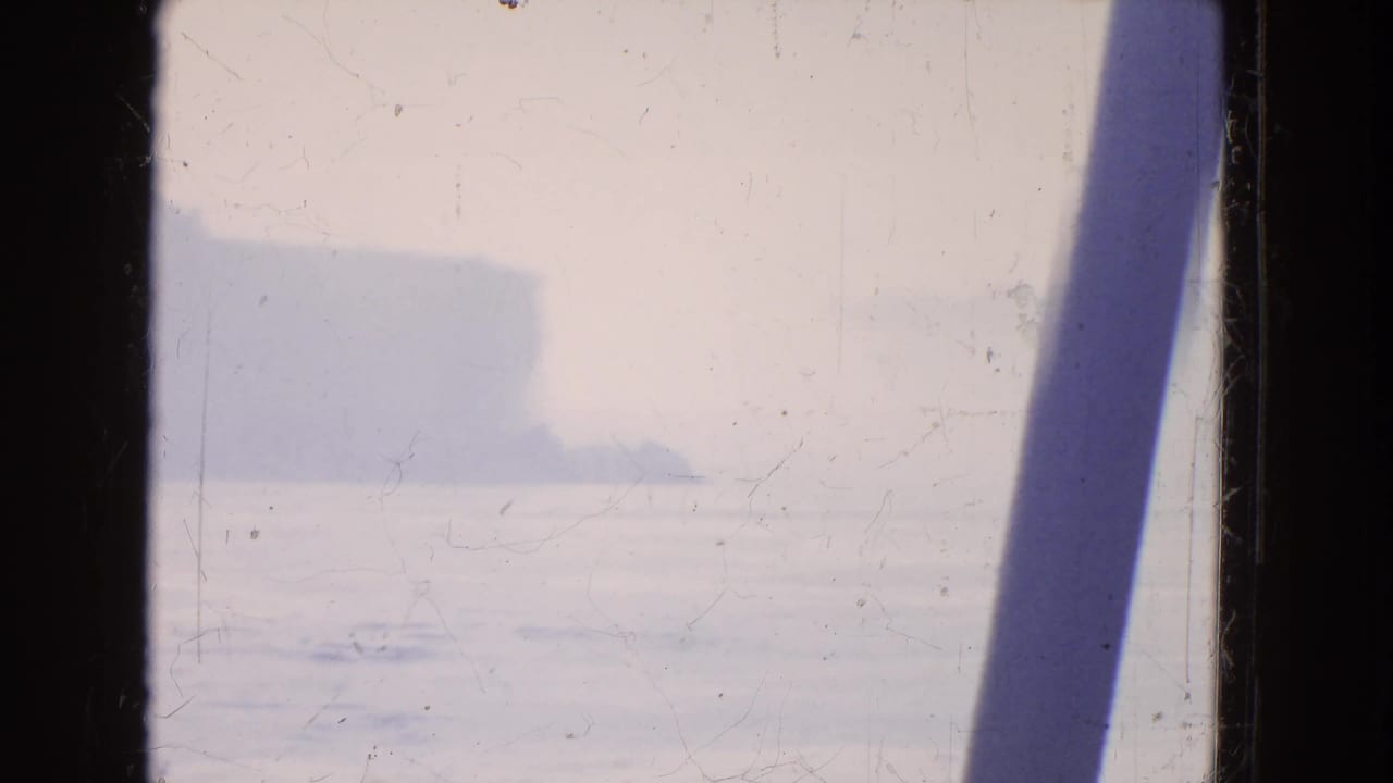 Faded 1964 8mm boat-view frame showing bright water, mist, distant shoreline, and a dark diagonal structural edge.