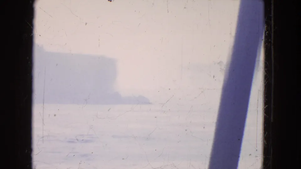 Faded 1964 8mm boat-view frame showing bright water, mist, distant shoreline, and a dark diagonal structural edge.