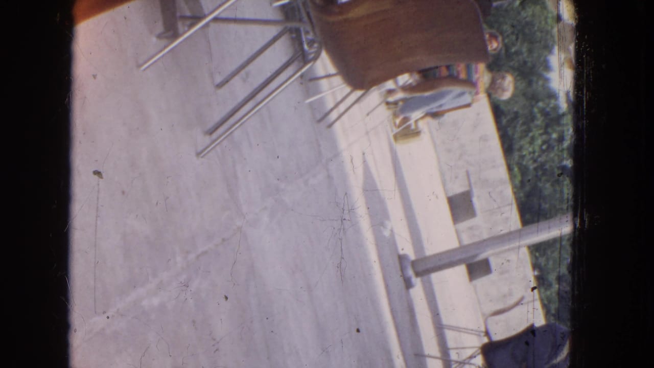 Tilted 8mm frame of concrete paving, metal chairs, a table, trees, and partially visible people.