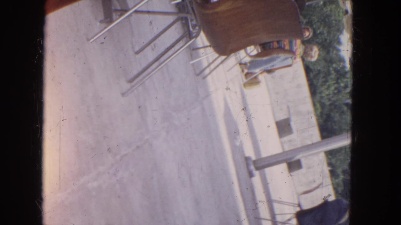 1964 Niagara Falls home-movie frame showing the same tilted outdoor seating area with metal chairs, concrete ground, trees, and cropped seated figures.
