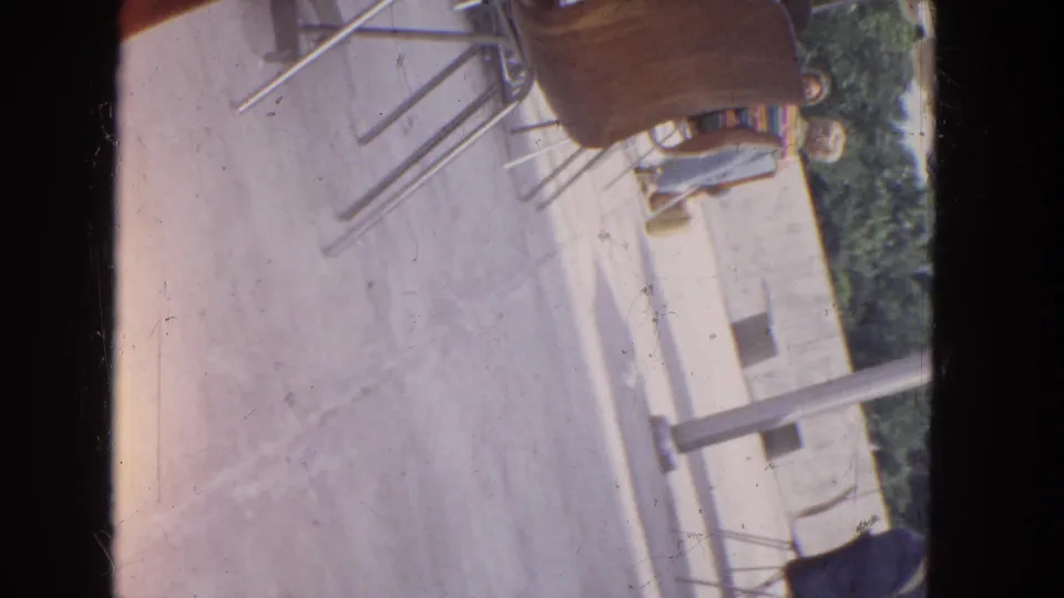 1964 Niagara Falls home-movie frame showing the same tilted outdoor seating area with metal chairs, concrete ground, trees, and cropped seated figures.