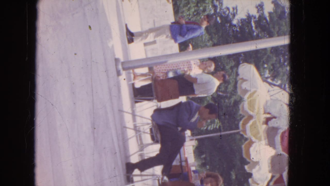 1964 Niagara Falls 8mm frame showing adults gathered near tables under a shade structure, with metal chairs, posts, trees, and concrete paving.