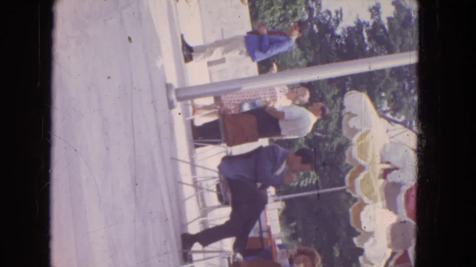 1964 Niagara Falls 8mm frame showing adults gathered near tables under a shade structure, with metal chairs, posts, trees, and concrete paving.