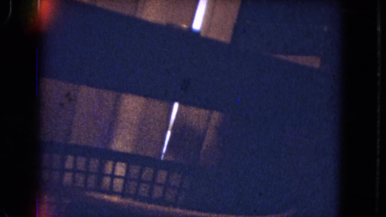 Low-light 1964 8mm view of a wooden exterior or structure, with diagonal dark bands, small light slits, and heavy blue grain.