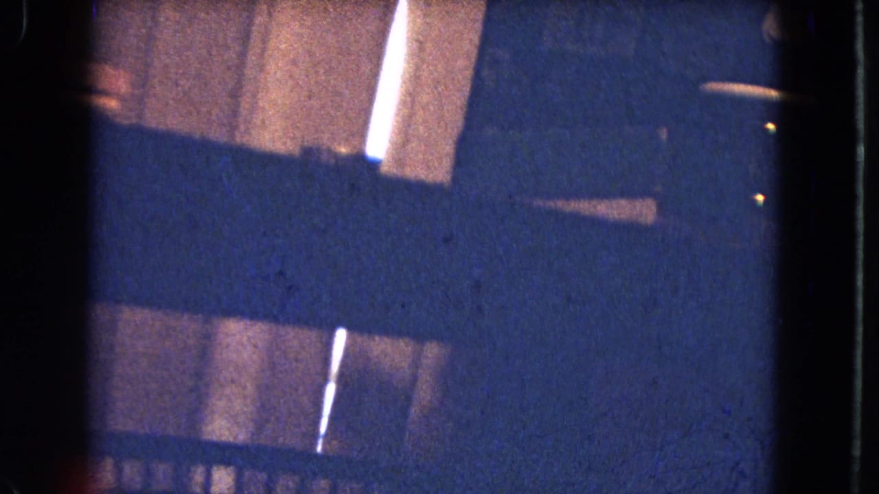 1964 8mm night frame showing a dim wooden structure or cabin exterior with dark diagonal obstruction, light slits, and visible film grain.