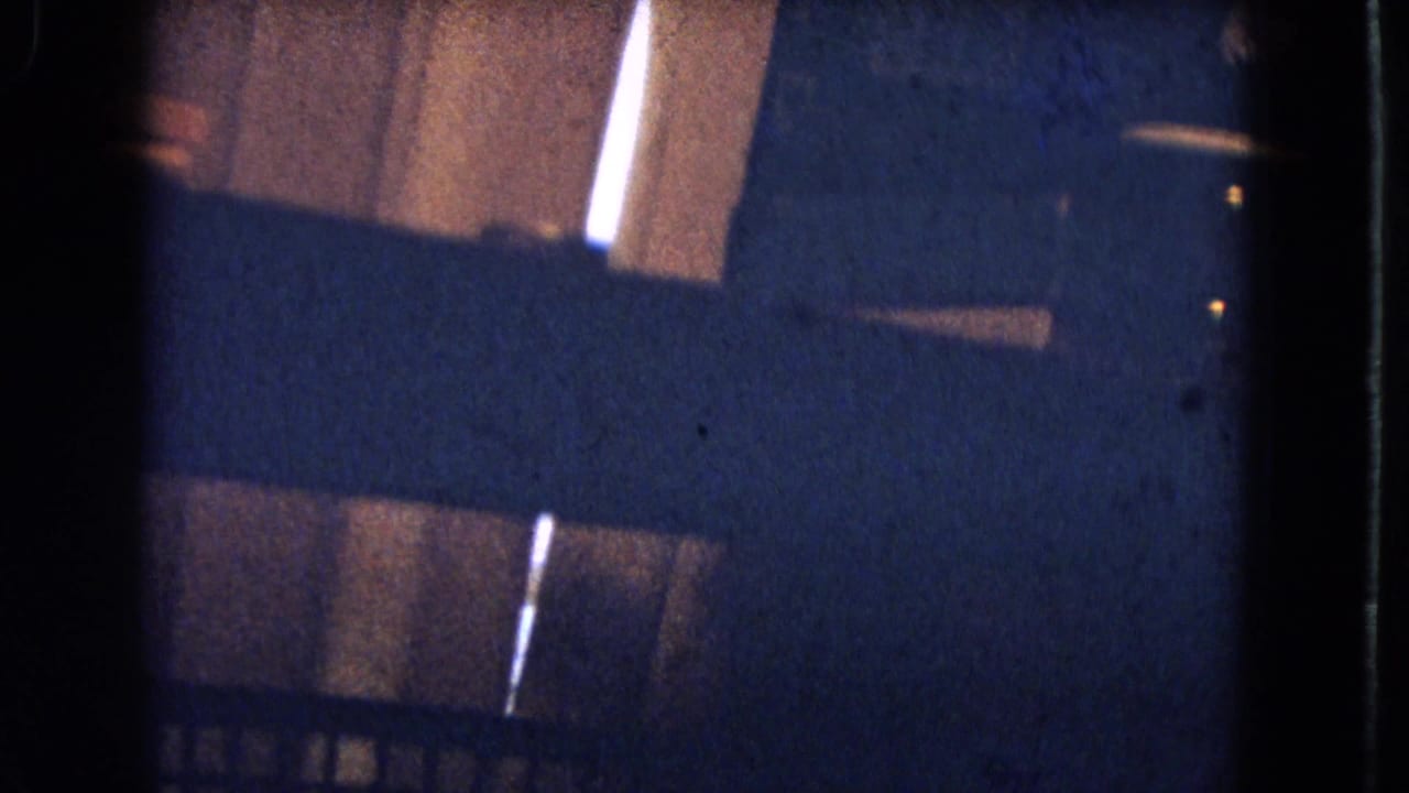 Dim interior frame with bed covers and part of a patterned wall, partly obscured by dark film edges.