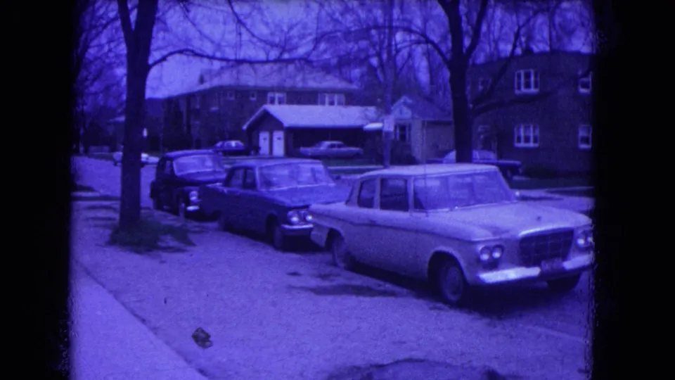 1964 Niagara Falls suburban street view with parked mid-century cars, brick houses, a sidewalk, and bare trees under a purple film cast.