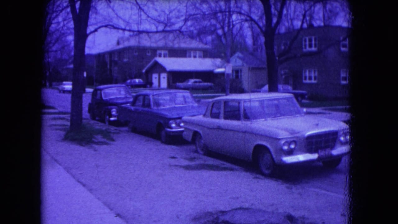 1964 Niagara Falls frame showing a residential block with parked cars, a sidewalk, lawns, bare trees, and neighboring houses.