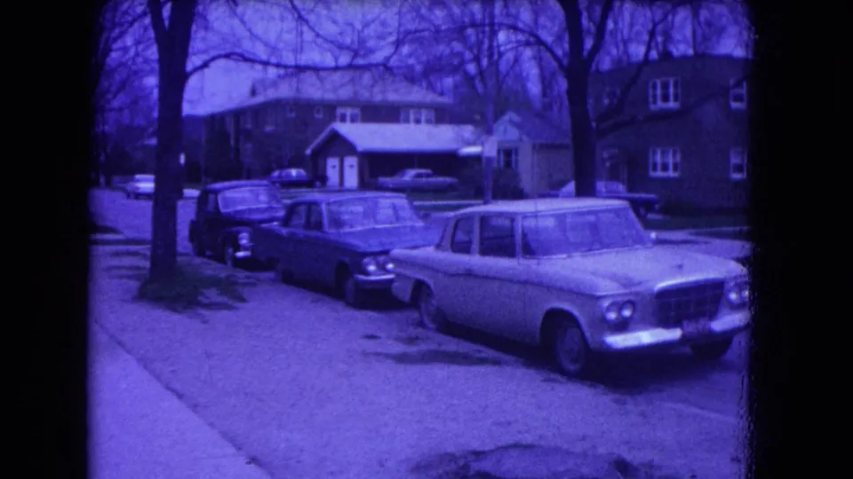 1964 Niagara Falls frame showing a residential block with parked cars, a sidewalk, lawns, bare trees, and neighboring houses.