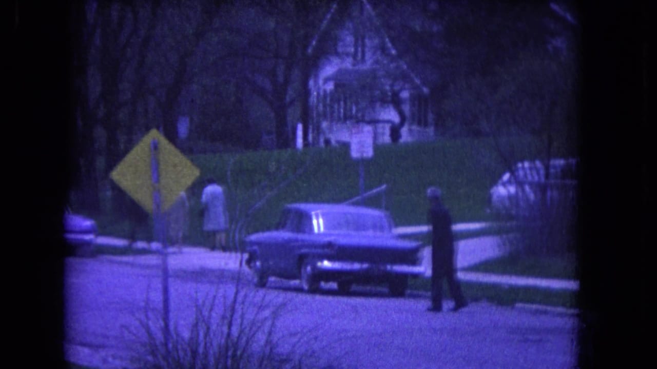 1964 Niagara Falls street scene with a parked classic car, yellow road sign, pedestrians, hedges, and a house in the background.