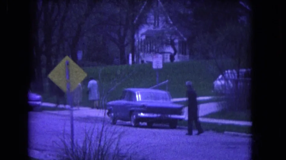 1964 Niagara Falls street scene with a parked classic car, yellow road sign, pedestrians, hedges, and a house in the background.