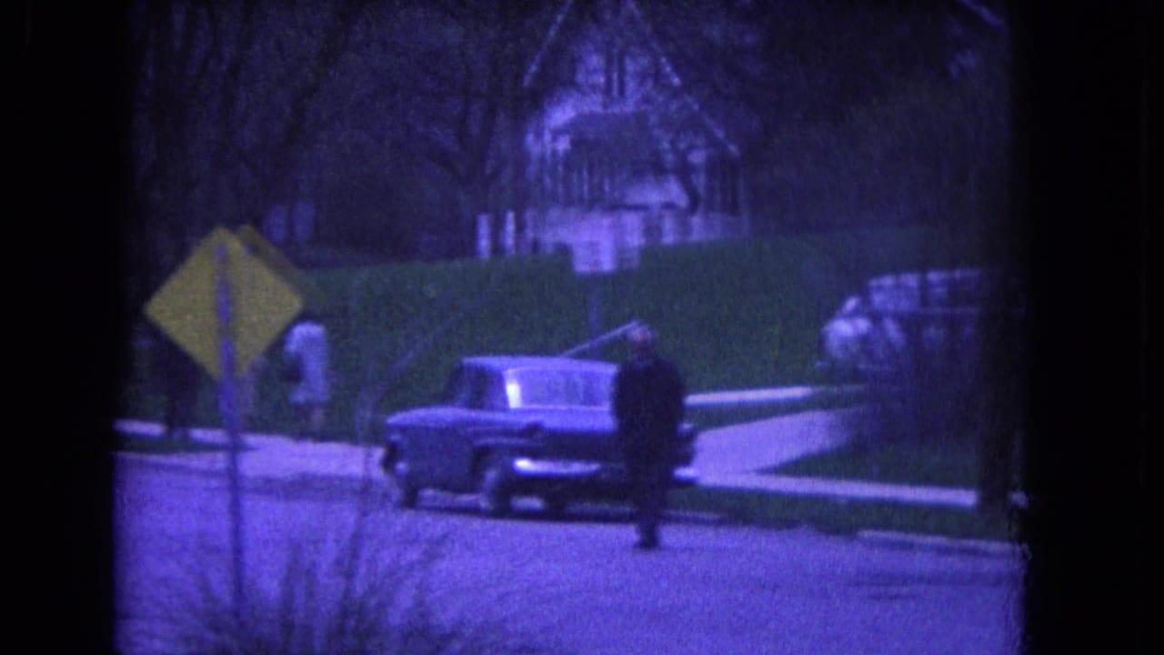 Niagara Falls, USA home-movie frame from 1964 showing a pedestrian, parked classic car, yellow sign, hedge, and residential backdrop.