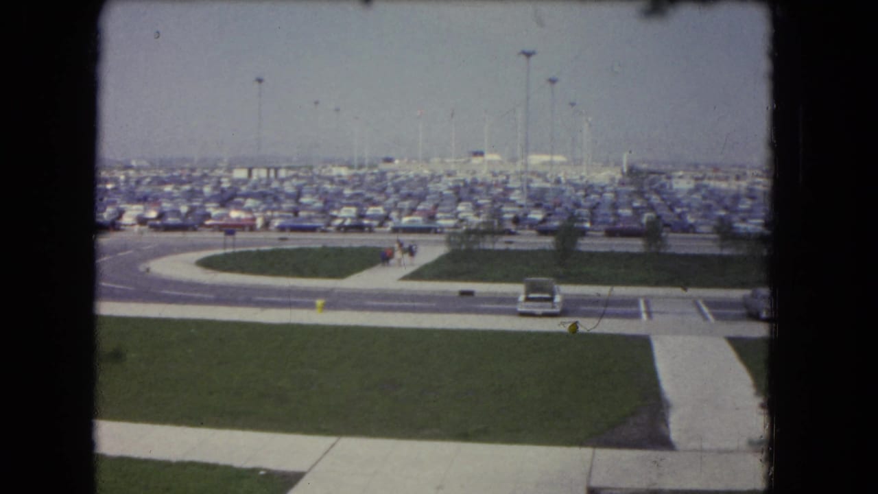 Wide parking lot filled with cars behind a curved road, sidewalks, lawns, and a few distant pedestrians.