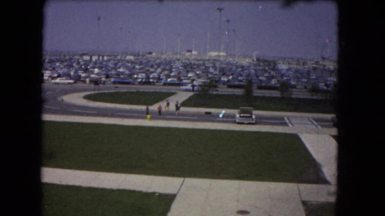 1964 8mm racetrack-area parking view with rows of cars, a curved entrance road, pedestrians, grass, and tall light poles.