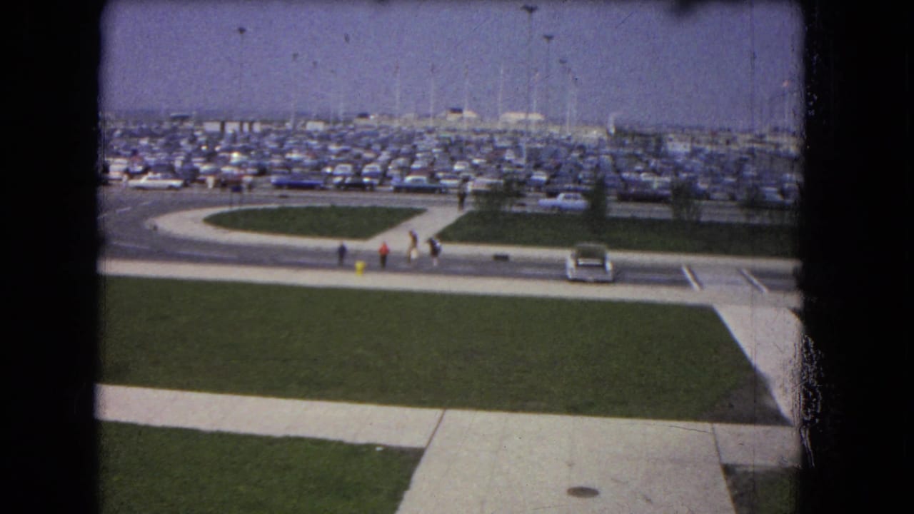 1964 8mm home-movie frame showing a crowded Niagara Falls-area event parking lot, sidewalks, grass, cars, and distant walkers.