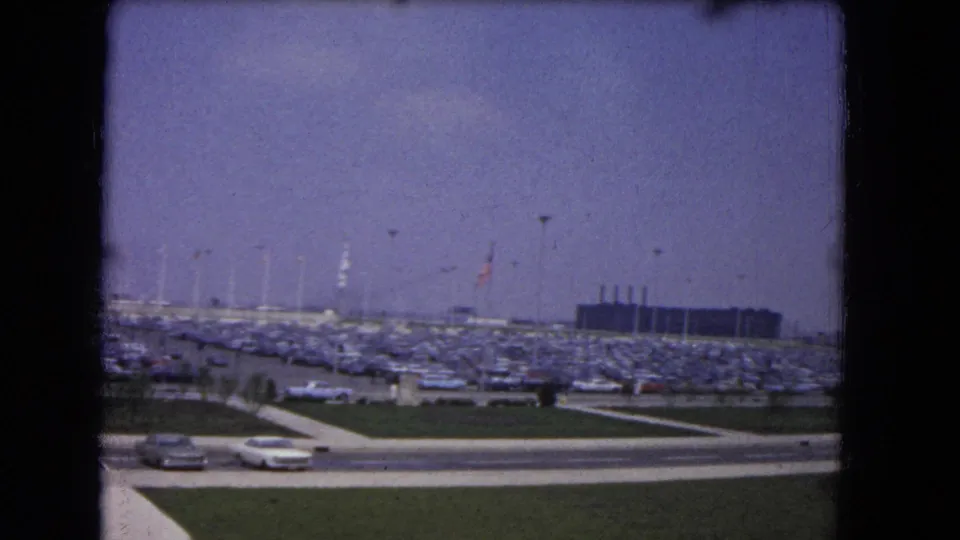 1964 8mm view across a crowded event parking lot with open sky, passing cars, grass strip, light poles, and a dark structure.