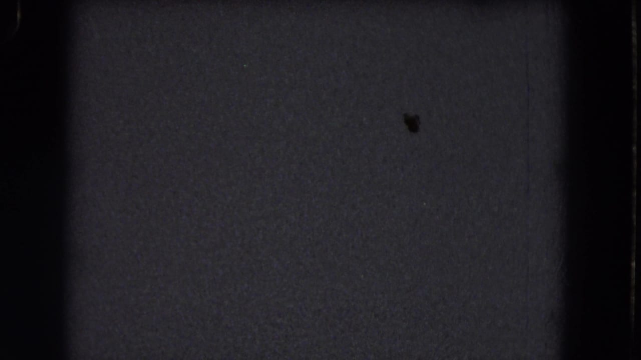 Dark 1964 8mm home-movie frame from the Niagara Falls reel showing grain, gate shadow, and a tiny dust mark.