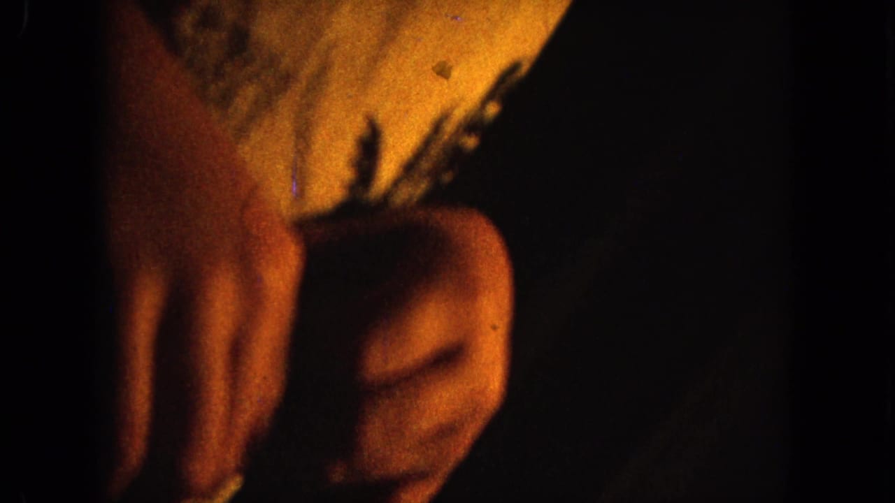 Dim 1964 Hawaii 8mm close-up of hands near a light patterned sleeve, framed tightly in warm interior light.