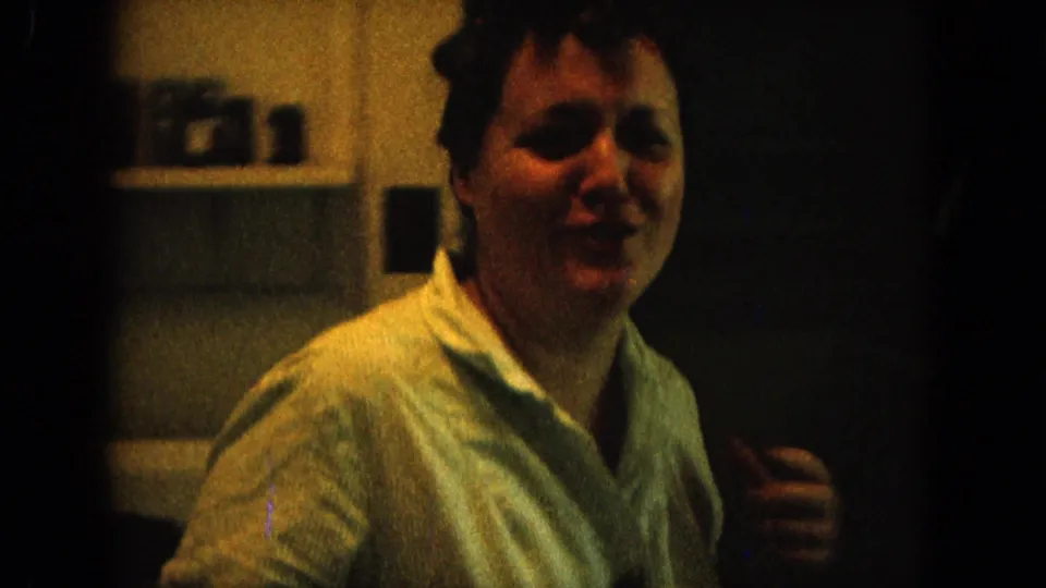 Young woman in a light collared shirt smiles in a dim 1964 Hawaii home interior, with shelves visible behind her.