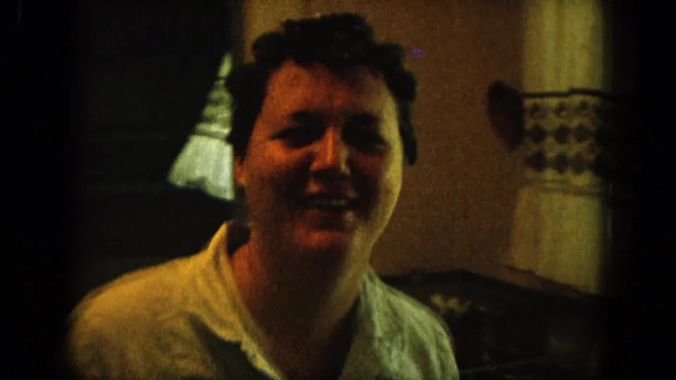 1964 Hawaii home-movie frame of a smiling woman with short dark hair in a light collared shirt inside a dim room.