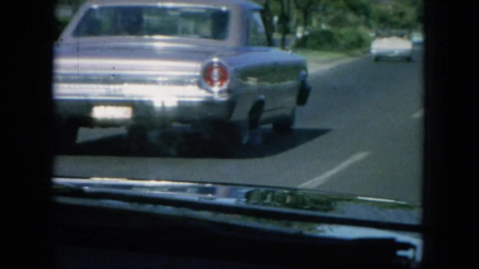 The camera follows a silver 1960s sedan on a paved coastal road, with another car dashboard visible in the foreground.
