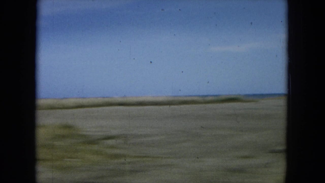 1964 Oahu 8mm frame showing a blurred coastal stretch from a car window, with sandy roadside, low horizon, and blue water beyond.