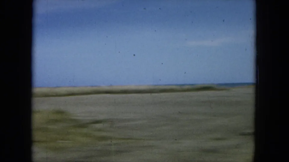 1964 Oahu 8mm frame showing a blurred coastal stretch from a car window, with sandy roadside, low horizon, and blue water beyond.