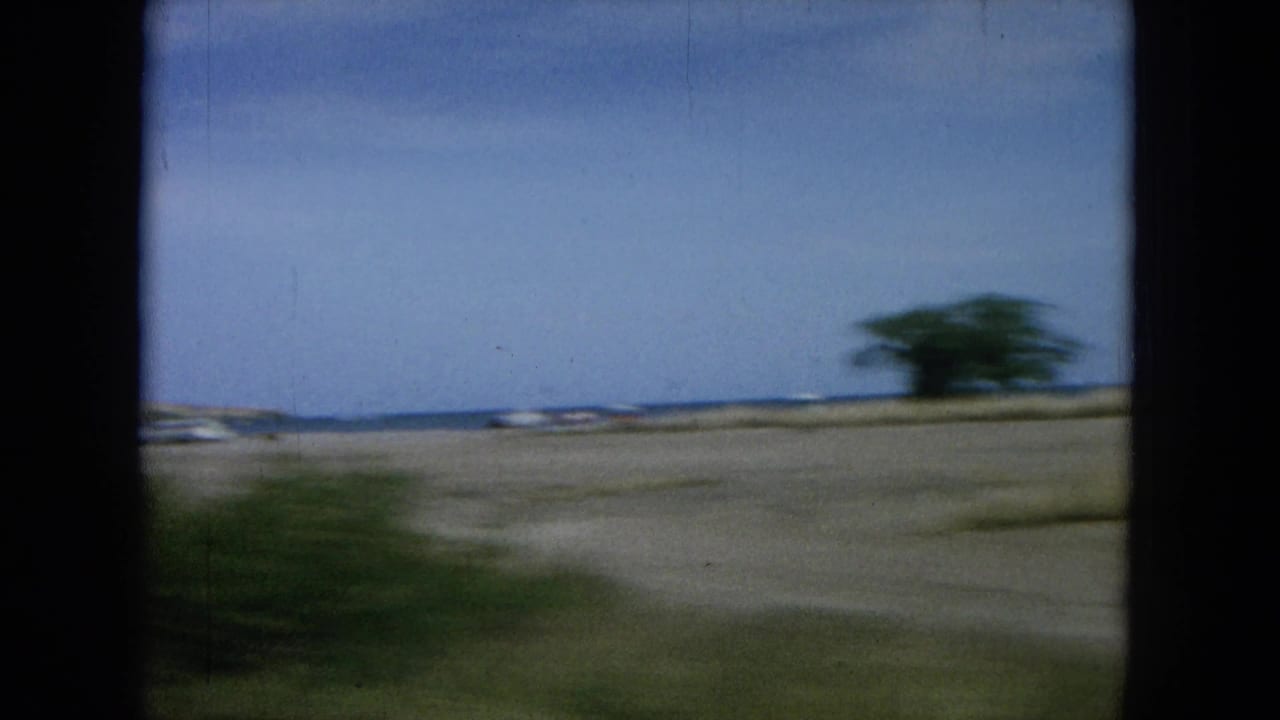 1964 Oahu home-movie frame from a moving vehicle, passing low coastal terrain, distant ocean, and roadside vegetation.