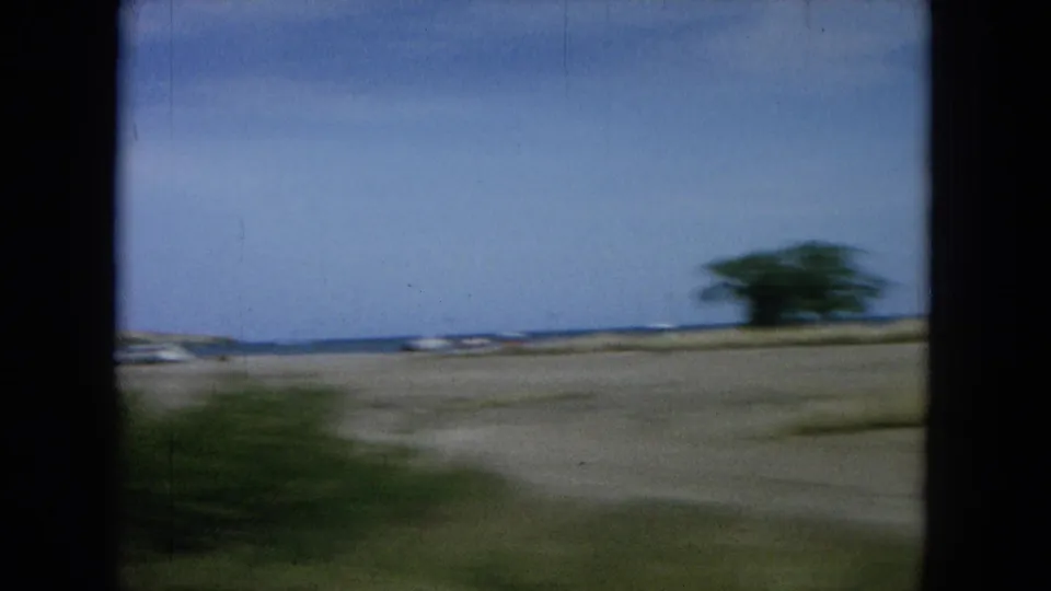 1964 Oahu home-movie frame from a moving vehicle, passing low coastal terrain, distant ocean, and roadside vegetation.