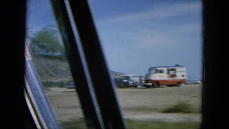 1964 Oahu 8mm frame looking from a vehicle toward parked mid-century cars and a small red-and-white camper near the coast.