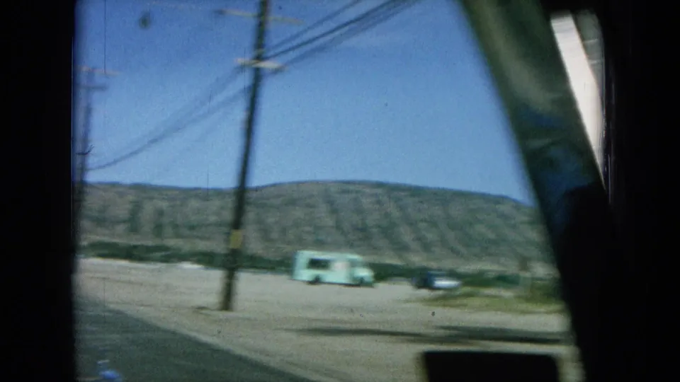 1964 Oahu 8mm frame from a moving car, showing utility wires, low hills, parked cars, and a pale green truck along the roadside.