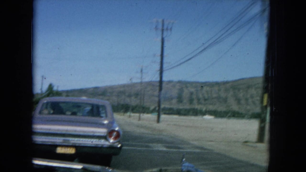 1964 8mm footage shows a car ahead on a sunny Oahu coastal road with utility poles and low hills.