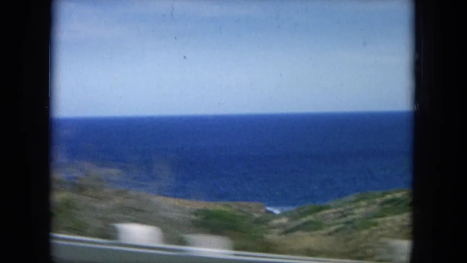 A soft 8mm view of the Oahu coast shows blue ocean, rocky shoreline, green slopes, and a white roadside rail.
