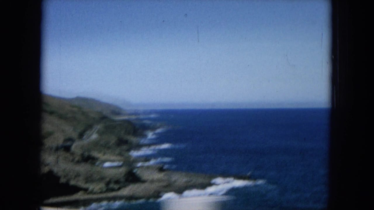Coastal hills and blue ocean seen from a moving roadside viewpoint.