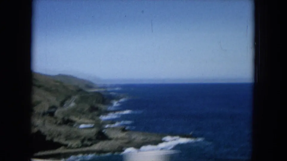 1964 Hawaii coastal drive view with rugged hills, blue ocean, surf lines, and pale sky seen from a moving vehicle.