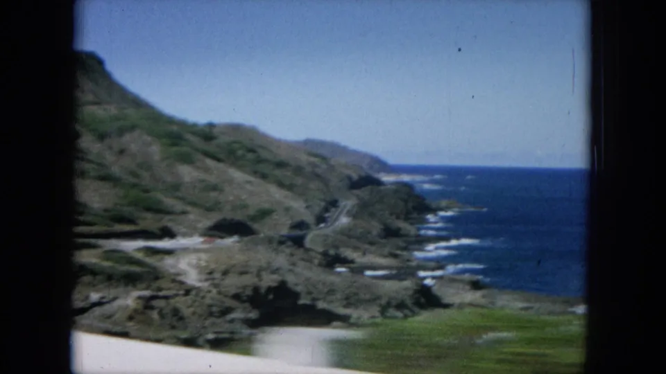 1964 Hawaii travel frame showing a hillside road perspective over rugged coast, blue sea, and white surf.