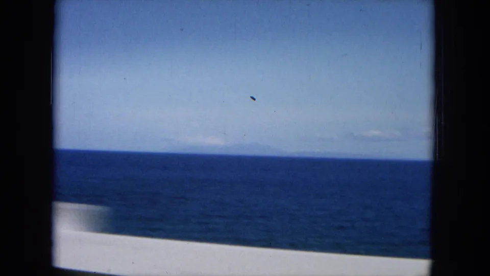 1964 Hawaii home-movie frame of open ocean, distant horizon, pale sky, and a blurred white roadside railing.