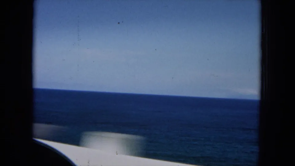 1964 Hawaii road-travel view of blue ocean and sky beyond a pale car body, recorded on grainy 8mm film.