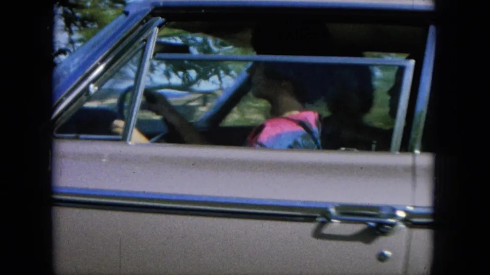 1964 Hawaii home-movie frame showing passengers inside a 1960s sedan, with roadside greenery behind the car.