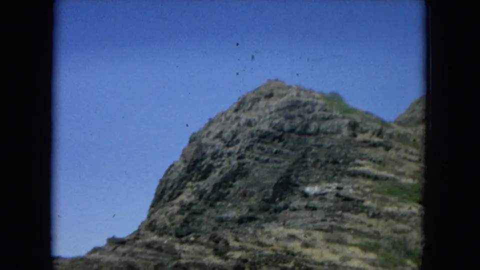 1964 Hawaii 8mm frame of a rugged mountain cliff rising under clear blue sky, with visible film grain and gate edge.