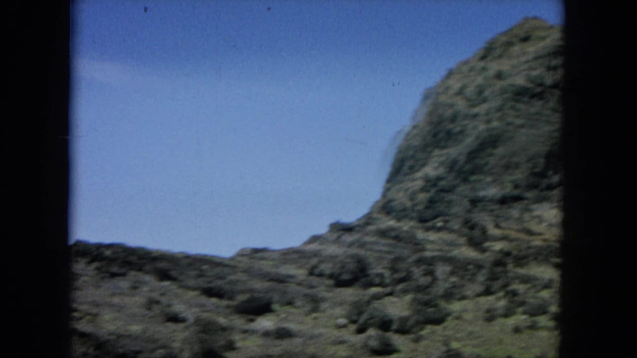 1964 8mm home-movie frame of a rugged Hawaiian coastal slope beneath a clear blue sky with dark projector borders.