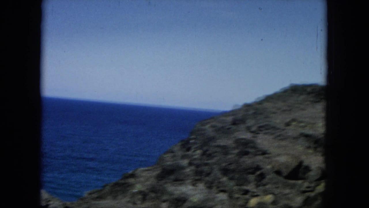 Ocean visible between rocky coastal slopes and dark vertical film borders.