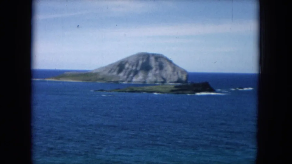 1964 8mm Hawaiian coastal view with a distant rocky island, surrounding blue water, and visible projector gate borders.