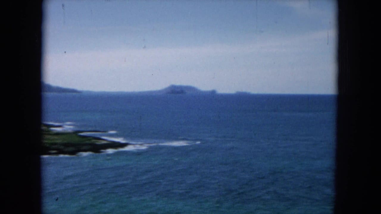 Open ocean with distant low islands and surf along a dark shoreline.