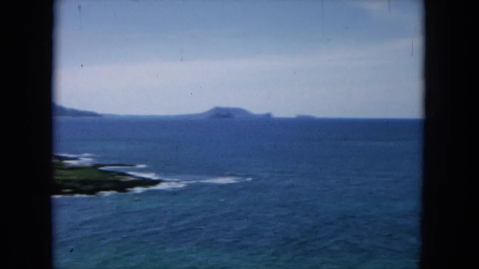 1964 8mm view from the Hawaiian coast showing deep blue sea, distant landforms, surf lines, and window-frame edges.